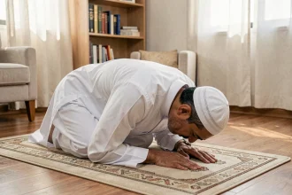 Homme en prière islamique en position de sujud sur un tapis de prière beige dans un salon simple au lever du soleil